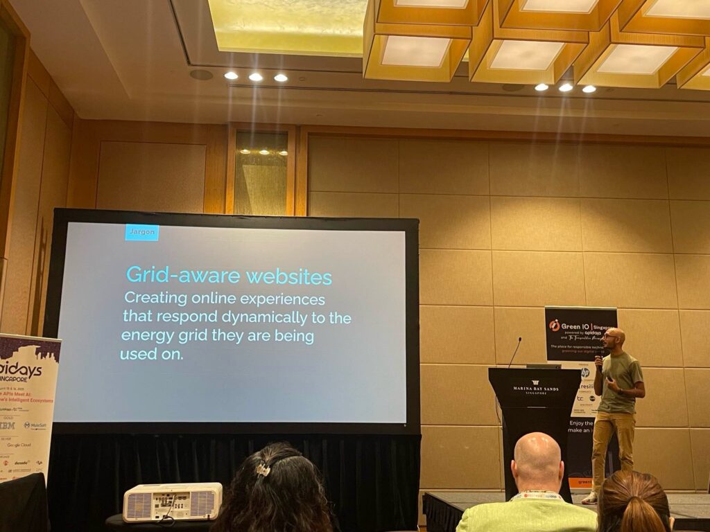 Text on a projector screen reading "Grid-aware websites - Creating online experiences that respond dynamically to the energy grid they are being used on." A bald man stands to the right of the screen next to a lectern, microphone in hand presenting the content.