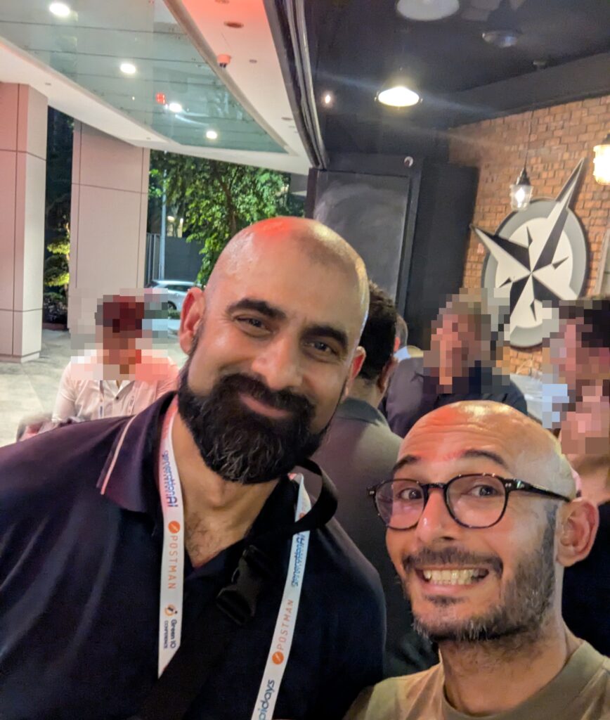 Two extremely good looking bald gentlemen posing for a selfie. They are in a outdoor bar setting, with other individuals in the background. The man on the left is taller, with a full black beard and slight smile. The man on the right is shorter, wearing black rimmed glasses.
