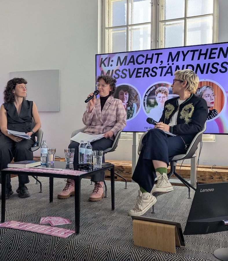 Three people are sitting on a stage, one person speaking in a microphone, the other two looking that direction. They are sitting in front of a screen with the text KI: Macht, Mythen, Missverständniss (the rest is not on the picture)